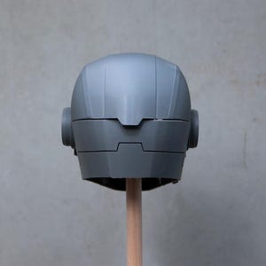 May include: A gray, futuristic-looking helmet with rounded ear pieces and a segmented design. The helmet is mounted on a wooden dowel against a plain gray background. The helmet's surface has a matte finish.