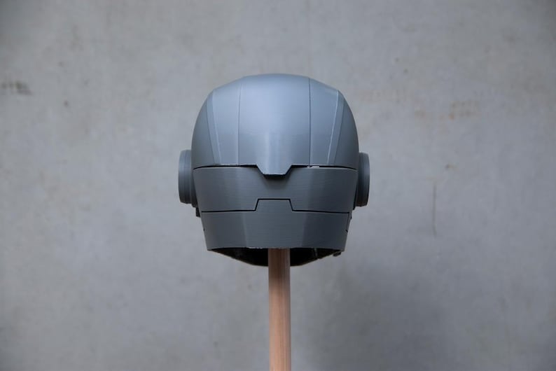 May include: A gray, futuristic-looking helmet with rounded ear pieces and a segmented design. The helmet is mounted on a wooden dowel against a plain gray background. The helmet's surface has a matte finish.