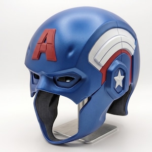 Op de afbeelding: Een blauwe en rode Captain America helm met een witte ster en de letter "A". De helm heeft een metallic afwerking en wordt gepresenteerd op een zilveren standaard. Het ontwerp bevat witte en rode accenten.