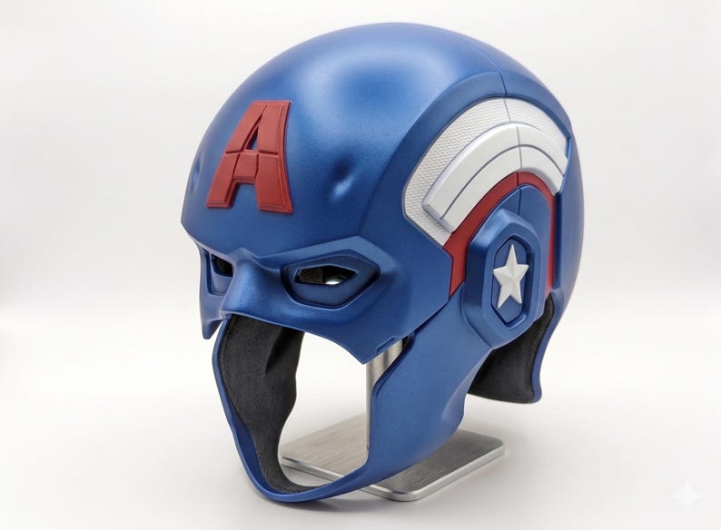 Op de afbeelding: Een blauwe en rode Captain America helm met een witte ster en de letter "A". De helm heeft een metallic afwerking en wordt gepresenteerd op een zilveren standaard. Het ontwerp bevat witte en rode accenten.