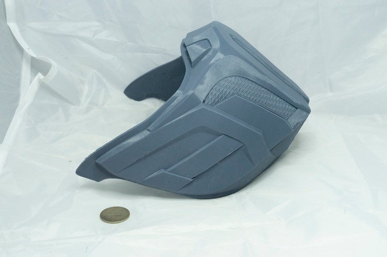 May include: A gray plastic mask with a textured surface and geometric designs. The mask is shaped like a half-face mask and has a curved shape.