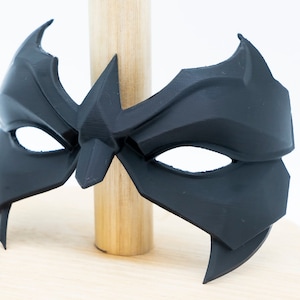 May include: A black plastic mask with a sharp, angular design. The mask has two eye holes and a pointed nose.
