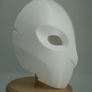 Op de afbeelding: Een witte, 3D-geprinte masker met een gladde afwerking. Het masker heeft een grote, ovale oogopening en een gebeeldhouwd ontwerp. Het is gemonteerd op een houten standaard, wat duidt op een display- of kostuumstuk.