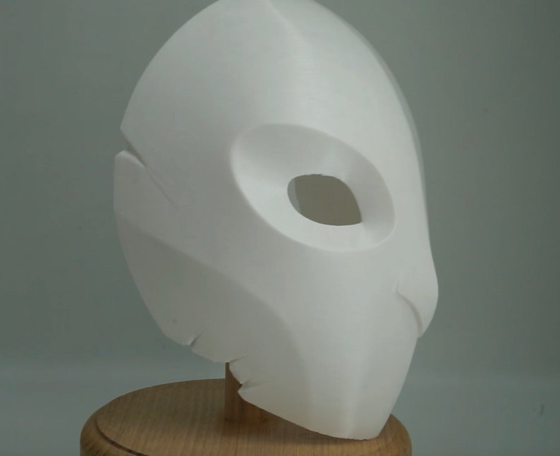 Op de afbeelding: Een witte, 3D-geprinte masker met een gladde afwerking. Het masker heeft een grote, ovale oogopening en een gebeeldhouwd ontwerp. Het is gemonteerd op een houten standaard, wat duidt op een display- of kostuumstuk.