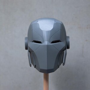 May include: A gray, geometric helmet with a futuristic design. The helmet features angular lines, eye openings, and side protrusions. It is mounted on a wooden dowel against a neutral background. The overall aesthetic suggests a sci-fi or cosplay theme.