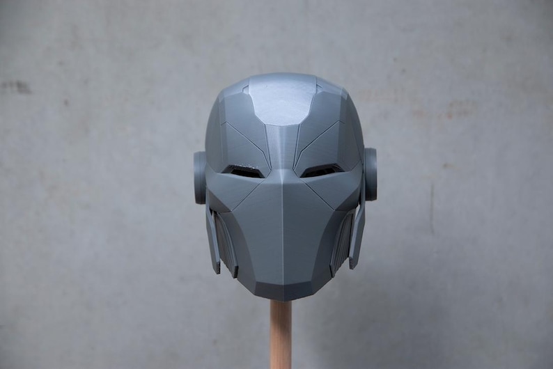 May include: A gray, geometric helmet with a futuristic design. The helmet features angular lines, eye openings, and side protrusions. It is mounted on a wooden dowel against a neutral background. The overall aesthetic suggests a sci-fi or cosplay theme.