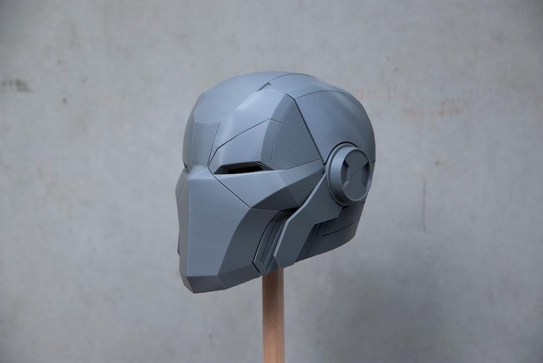 May include: A gray, futuristic-style helmet with angular panels and a circular detail on the side. The helmet is mounted on a wooden dowel, showcasing its design and structure. The overall aesthetic suggests a sci-fi or cosplay theme.