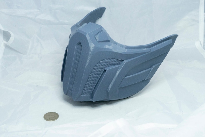 May include: A gray plastic 3D printed mask with a textured surface. The mask has a sharp, angular design with a pointed top and a wide, flat bottom.