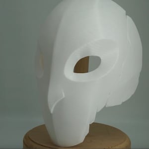 Op de afbeelding: Een witte, 3D-geprinte masker met grote, ovale ooggaten en een glad, gebogen oppervlak. Het masker heeft een gestileerd, vogelachtig ontwerp, mogelijk voor cosplay of kostuums. Het wordt gepresenteerd op een houten standaard tegen een neutrale achtergrond.