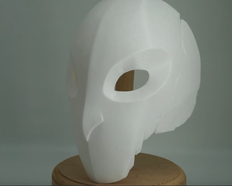 Op de afbeelding: Een witte, 3D-geprinte masker met grote, ovale ooggaten en een glad, gebogen oppervlak. Het masker heeft een gestileerd, vogelachtig ontwerp, mogelijk voor cosplay of kostuums. Het wordt gepresenteerd op een houten standaard tegen een neutrale achtergrond.