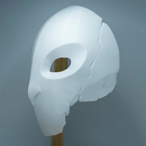 Op de afbeelding: Een wit, gebeeldhouwd masker met een ovale oogopening en een gedeeltelijk gebroken oppervlak. Het masker is gemonteerd op een houten deuvel tegen een grijze achtergrond, wat duidt op een rekwisiet of kostuumelement.