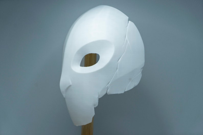 Op de afbeelding: Een wit, gebeeldhouwd masker met een ovale oogopening en een gedeeltelijk gebroken oppervlak. Het masker is gemonteerd op een houten deuvel tegen een grijze achtergrond, wat duidt op een rekwisiet of kostuumelement.
