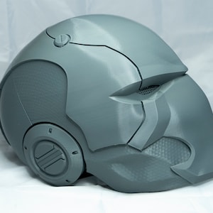 May include: A gray 3D printed helmet with a textured surface and a circular design on the side. The helmet has a visor and a small detail on the top.