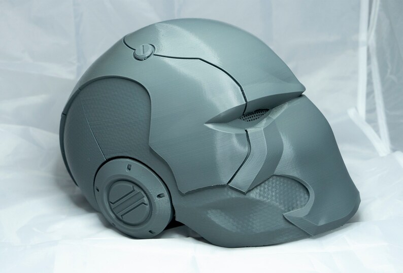 May include: A gray 3D printed helmet with a textured surface and a circular design on the side. The helmet has a visor and a small detail on the top.