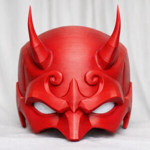 May include: A bright red Oni mask featuring two horns, detailed facial features, and mesh eye coverings. This 3D-printed mask embodies a traditional Japanese demon design, ideal for cosplay or display.