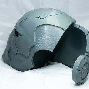 May include: A gray 3D printed helmet with a textured surface and a removable side panel. The helmet has a circular detail on the side.