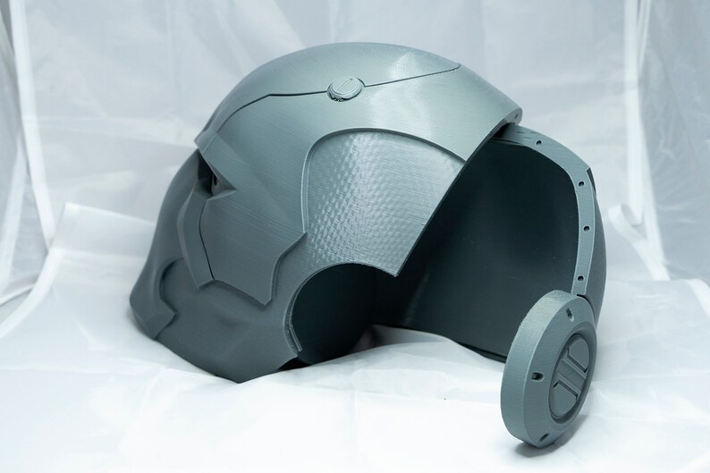 May include: A gray 3D printed helmet with a textured surface and a removable side panel. The helmet has a circular detail on the side.