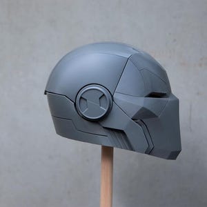 May include: A gray, futuristic-style helmet with angular design elements. The helmet features a circular emblem on the side and a face covering. It is mounted on a wooden dowel against a neutral background.