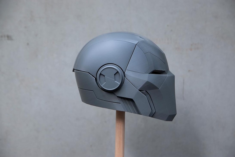 May include: A gray, futuristic-style helmet with angular design elements. The helmet features a circular emblem on the side and a face covering. It is mounted on a wooden dowel against a neutral background.