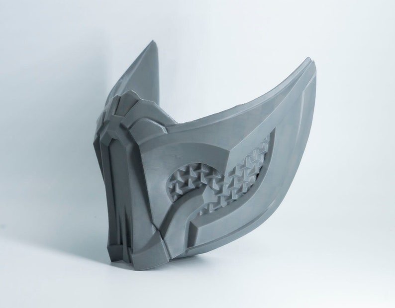 May include: A gray 3D printed mask with a geometric design. The mask has a pointed top and a curved bottom.
