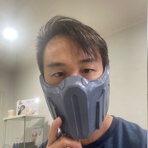 May include: A person wearing a gray, 3D printed mask that covers the nose and mouth. The mask has a geometric design with raised lines and a textured surface.