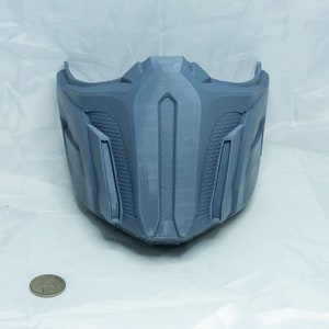 May include: A gray 3D printed mask with a detailed design. The mask has a pointed chin and a raised, vertical ridge in the center.