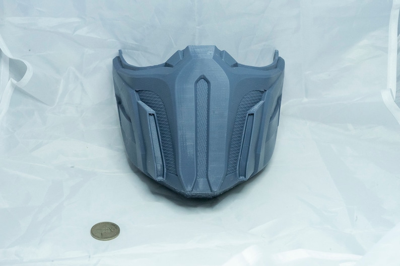 May include: A gray 3D printed mask with a detailed design. The mask has a pointed chin and a raised, vertical ridge in the center.