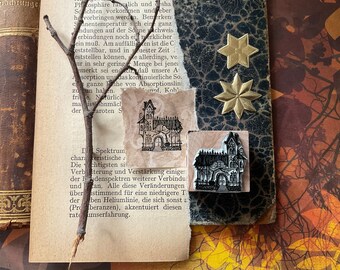 May include: Two black and white rubber stamps of a spooky house with a pointed roof, windows, and a door. The stamps are on a brown and white textured surface.