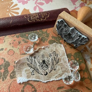 Könnte beinhalten: Ein Holzstempel mit einem Blumen- und Insektenmuster liegt neben einem zerrissenen Papierstück mit einer passenden Illustration. Klare, dekorative Knöpfe und ein Vintage-Buch sind ebenfalls im Bild. Der Hintergrund zeigt ein gemustertes Papier.