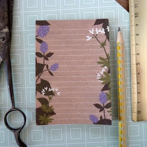 Könnte beinhalten: Ein braunes Notizbuch mit linierten Seiten und einem Blumendesign mit lila und weißen Blüten. Das Notizbuch liegt auf einer grün-weiß gemusterten Oberfläche mit einer Schere und einem Lineal.