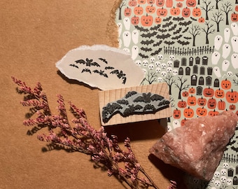 May include: A set of two Halloween-themed rubber stamps featuring bats. One stamp shows a group of bats flying in a cluster, while the other stamp shows a larger group of bats flying in a formation. The stamps are on a brown background with a pink rock and a piece of paper with a Halloween-themed pattern.