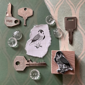 Könnte beinhalten: Eine flache Ansicht einer Sammlung von Vintage-Schlüsseln, Glasknöpfen und einem Gummibild eines Vogels. Der Stempel befindet sich auf einem Holzblock und die Schlüssel sind silberfarben mit einer abgenutzten Oberfläche. Die Knöpfe sind aus klarem Glas mit einem erhabenen Design.