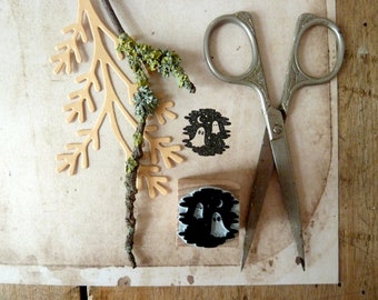 May include: Two wooden stamps with a black and white design of ghosts and a crescent moon. The stamps are on a beige surface with a brown twig and a pair of silver scissors.