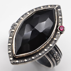 May include: A large, ornate ring featuring a marquise-cut black gemstone. The ring has a silver-toned setting with a textured border and a small, round red gemstone accent. The band has a detailed floral design.