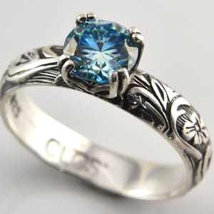 May include: A silver ring with a large, round, light blue gemstone. The band features an intricate floral and leaf design. The word "CUDS" is engraved on the inside of the ring.