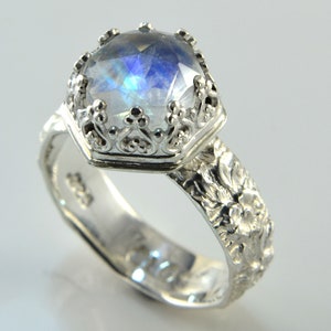 Op de afbeelding: Zilveren ring met een grote, iriserende edelsteen in een kroonachtige zetting. De band is versierd met bloemmotieven. Het ontwerp van de ring is elegant en geïnspireerd op de natuur.