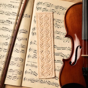 May include: A beige crocheted bookmark with a delicate lace pattern lies on a sheet of music with a violin and bow. The music sheet has the text "F minor" and "Melodic Scale" printed on it.