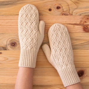 Könnte beinhalten: Ein Paar cremefarbene Strickfäustlinge mit Rautenmuster. Die Fäustlinge werden auf einer Holzoberfläche präsentiert. Sie haben gerippte Bündchen und ein strukturiertes Design, ideal für kaltes Wetter.