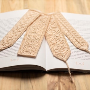 May include: Three beige crocheted bookmarks with intricate patterns, lying on an open book.