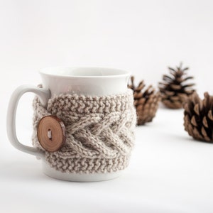 May include: White ceramic mug with a knitted beige cozy featuring a wooden button. The mug is surrounded by pine cones on a white background.