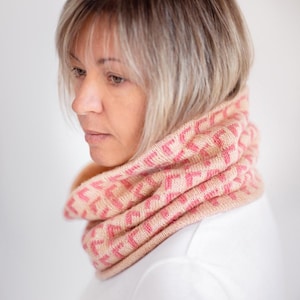 May include: A knitted cowl in shades of light pink and cream, featuring a geometric pattern. The cowl is wrapped around a person's neck, with the design visible. The person is wearing a white shirt. The background is a soft, neutral color.