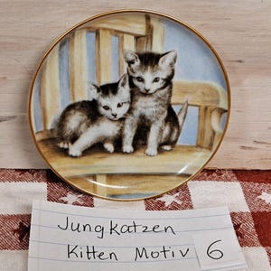 May include: A porcelain plate with a gold rim features a painting of two kittens sitting on a wooden chair. The plate has a light blue background and the kittens are brown and white.