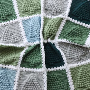 May include: A crocheted blanket with a geometric pattern of green, blue and grey squares. Each square features a raised, textured design resembling a tree. The blanket is edged with a white crocheted border.