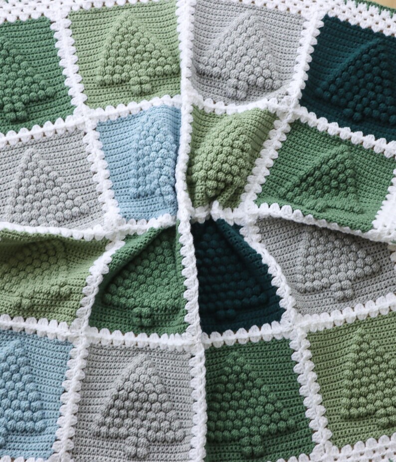 May include: A crocheted blanket with a geometric pattern of green, blue and grey squares. Each square features a raised, textured design resembling a tree. The blanket is edged with a white crocheted border.