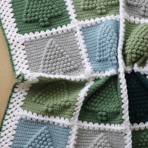 May include: A crocheted blanket with a patchwork design featuring squares in shades of green, blue, and grey. The squares are edged with a white crocheted border.