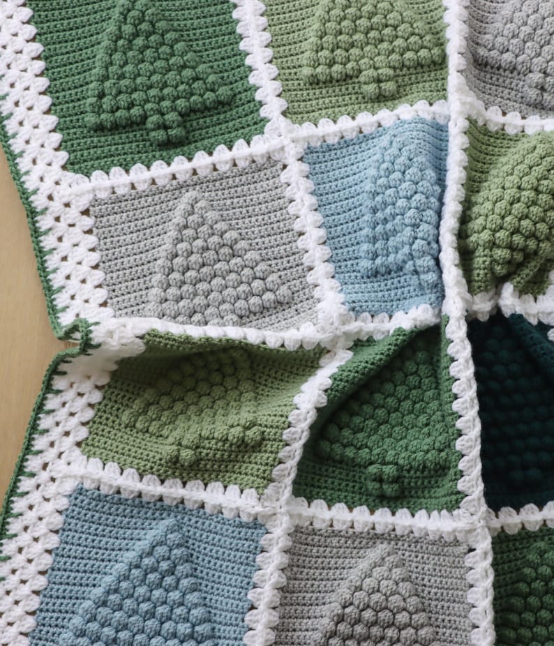 May include: A crocheted blanket with a patchwork design featuring squares in shades of green, blue, and grey. The squares are edged with a white crocheted border.