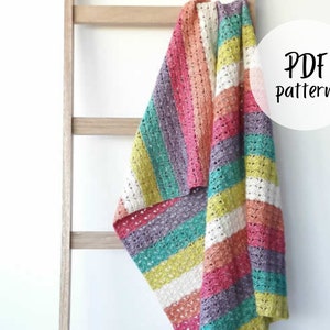 Op de afbeelding: Een kleurrijk gestreept gehaakt deken gedrapeerd over een houten ladder. Het deken is gemaakt met verschillende kleuren, waaronder roze, oranje, geel, groen en paars. De tekst "PDF Pattern" is geschreven in een witte cirkel aan de rechterkant van de afbeelding.
