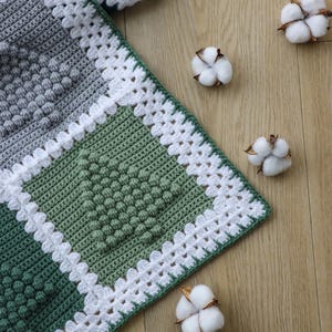 May include: A crocheted blanket with a white border and a green and grey pattern. The blanket is made with a textured stitch that creates a raised, bumpy effect. The blanket is lying on a wooden surface with cotton balls scattered around it.