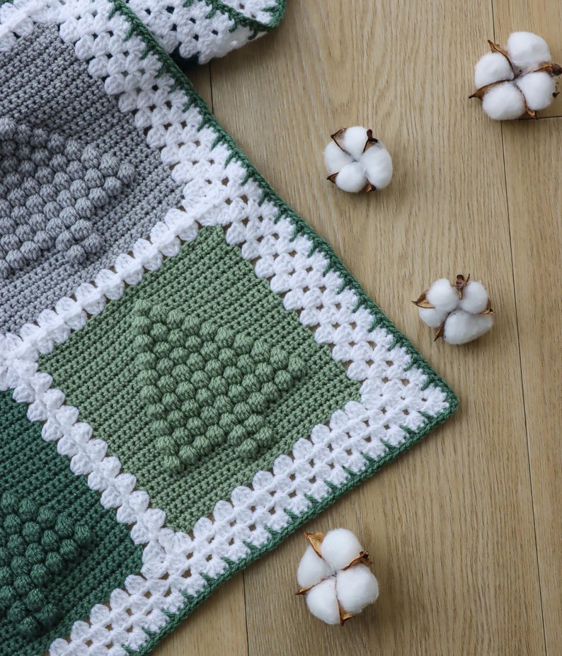 May include: A crocheted blanket with a white border and a green and grey pattern. The blanket is made with a textured stitch that creates a raised, bumpy effect. The blanket is lying on a wooden surface with cotton balls scattered around it.