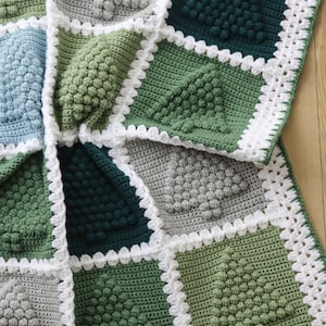 May include: A crocheted blanket with a white border and a geometric pattern of green, blue, and grey squares. The blanket is made with a textured stitch that creates a raised, bumpy surface.